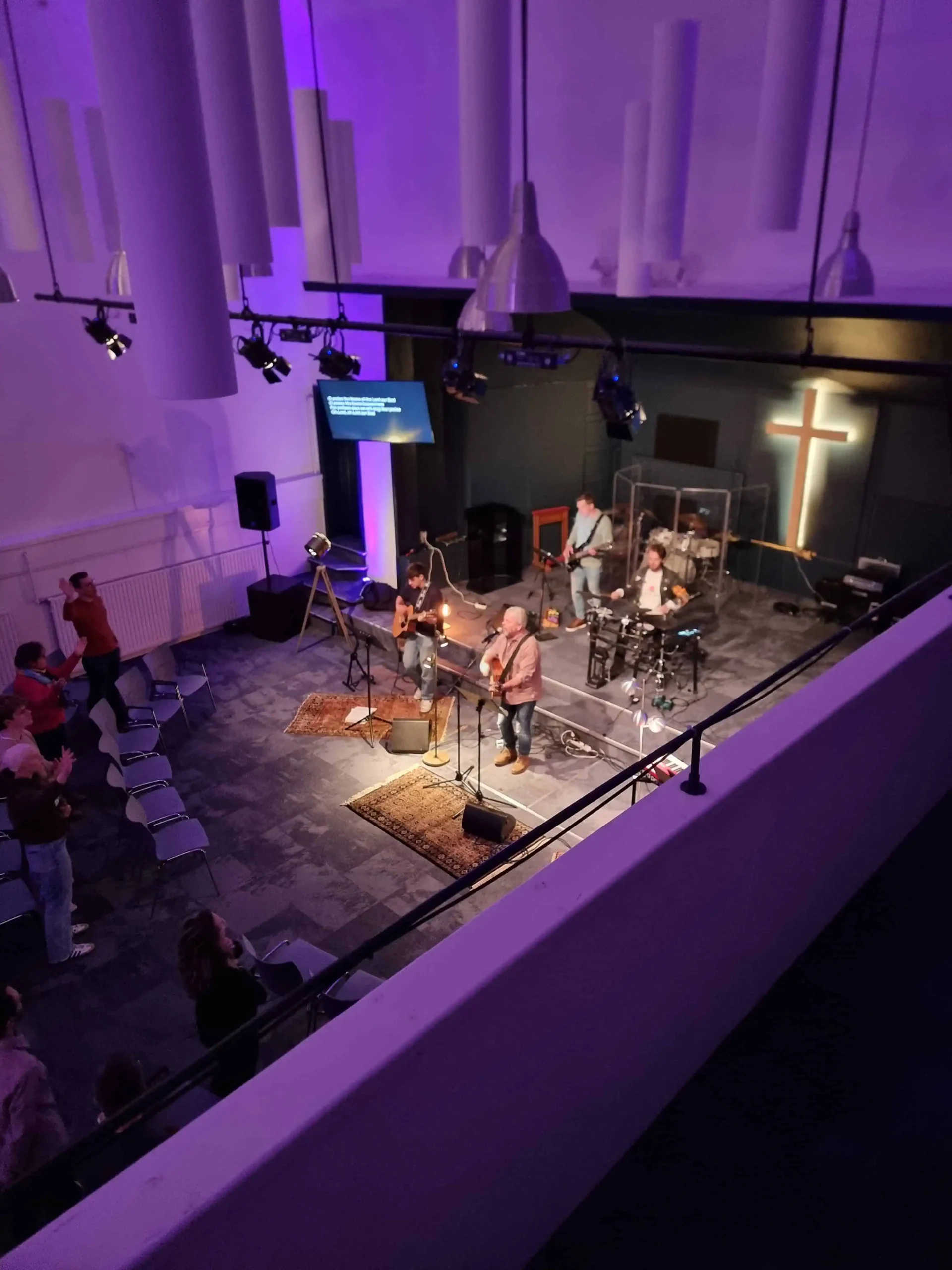 Muziekband speelt op een podium met een verlicht kruis en paarse belichting tijdens een worshipdienst, terwijl bezoekers staand meezingen.