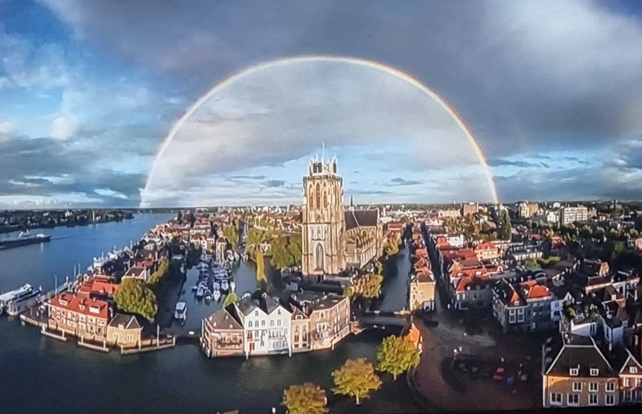 Luchtfoto van de historische binnenstad van Dordrecht met de Grote Kerk aan het water en een volledige regenboog boven de stad.