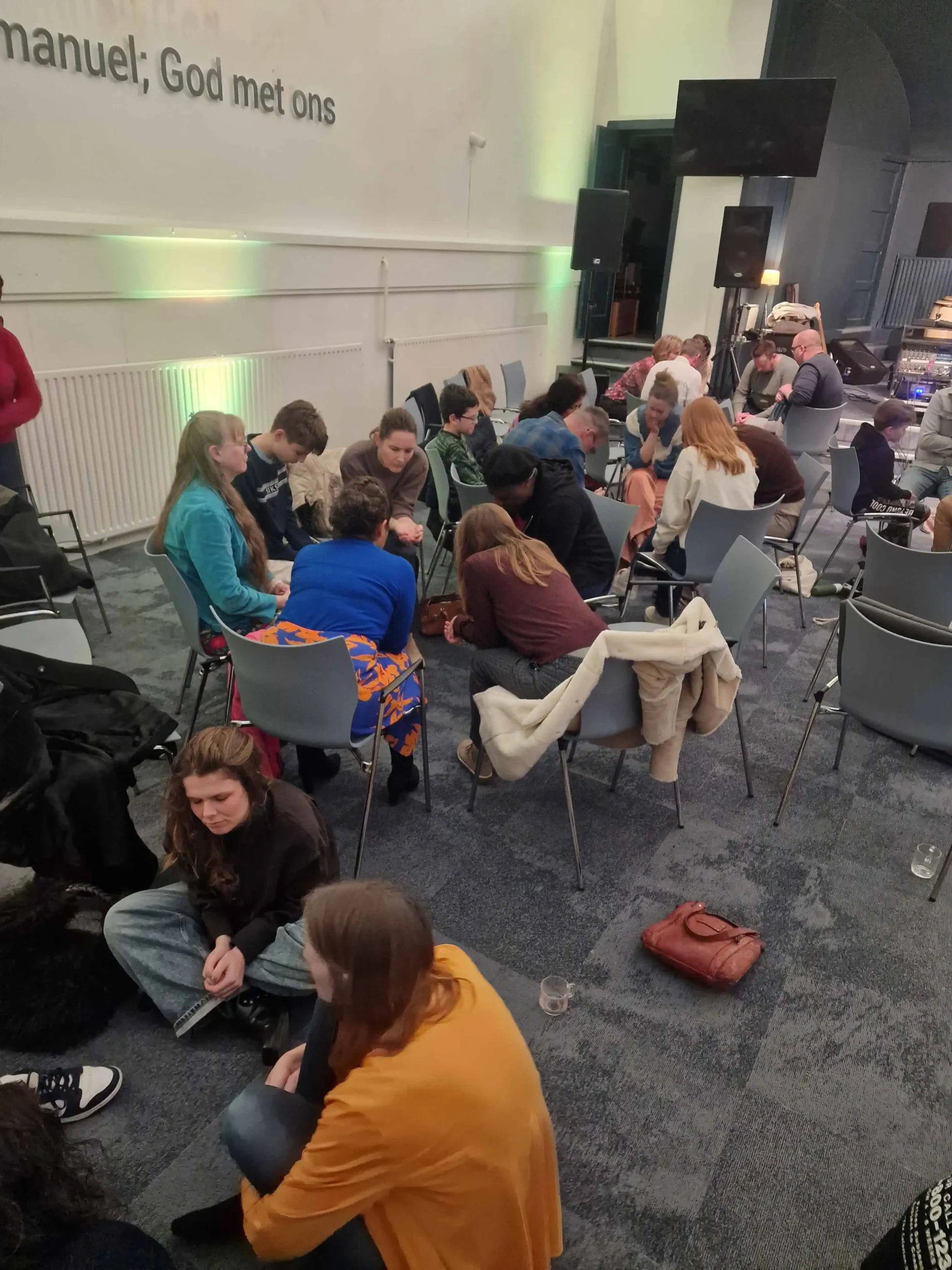 Groep mensen zit op stoelen en op de grond met gebogen hoofden te bidden in een zaal, met op de muur de tekst Immanuel, God met ons.