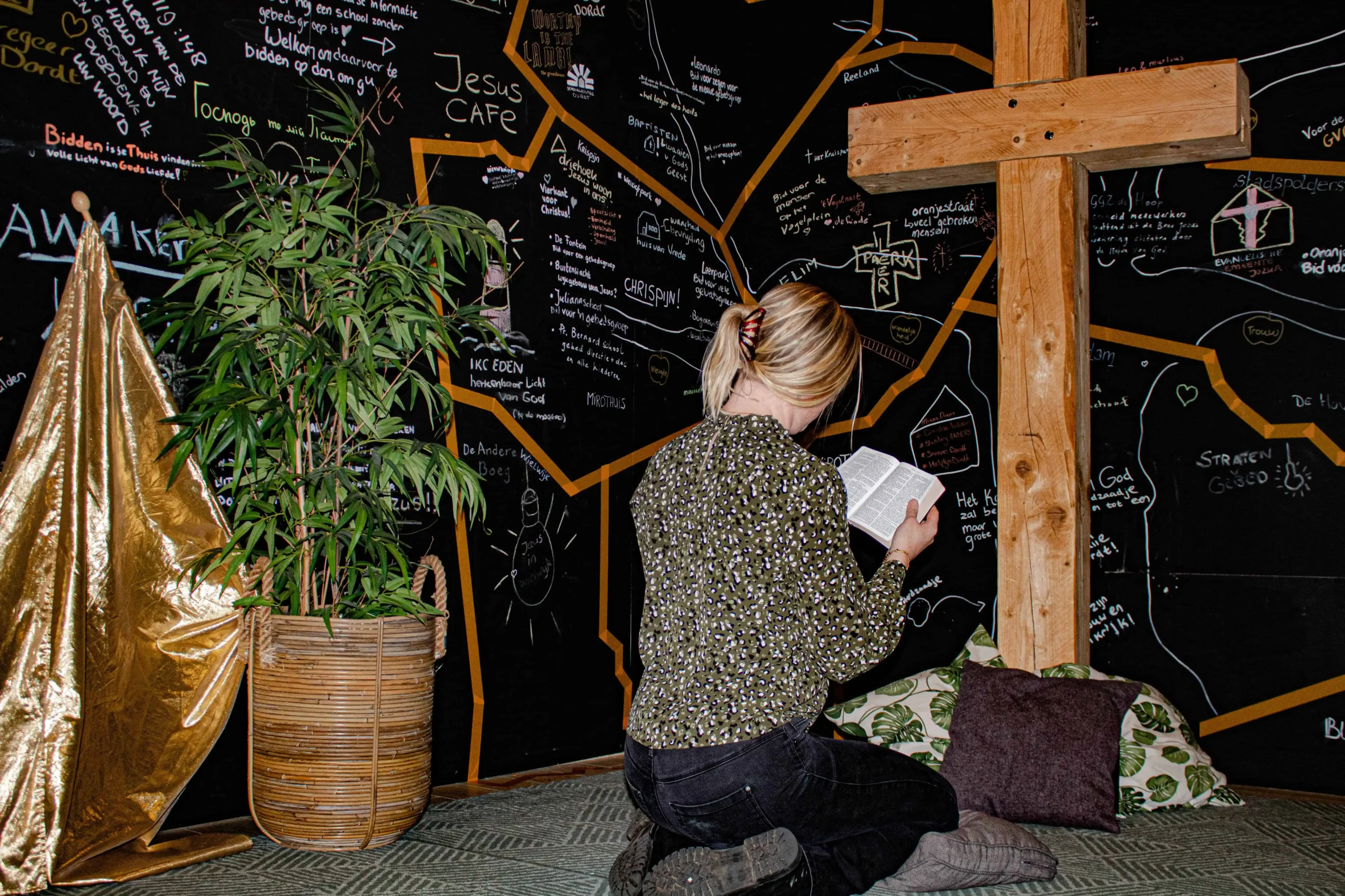 Vrouw knielt en leest in een Bijbel voor een groot houten kruis en een zwarte gebedsmuur vol krijtteksten, naast een plant en kussens.